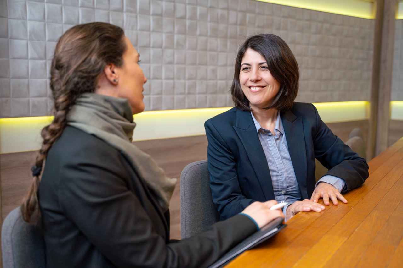 Eine Mitarbeiterin des Holiday Inn Munich City Centre der Abteilung Commercial Team führt ein Gespräch mit einer Frau unter vier Augen. Sie sitzen gemeinsam an einem Tisch und schauen sich in die Augen.